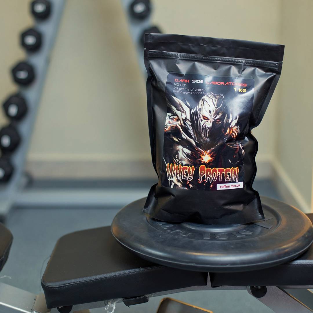 a pack of whey protein from Dark Side Laboratories, standing on a plate for a barbell, in the background, a dumbbell row of a gym